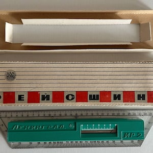 May include: A green plastic drafting tool with a clear ruler base, positioned below a cardboard box. The box features horizontal black lines, a small ship logo, and the Cyrillic text "РЕЙСШИНА" in black letters on a red background. The tool displays "ЛЕНИНГРАД" and "ИР-2".