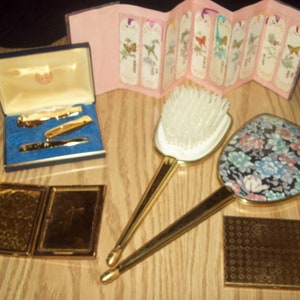 May include: A set of vintage gold-toned vanity items including a compact mirror, a hairbrush, a hand mirror with a floral pattern, and a small rectangular box with a patterned lid.