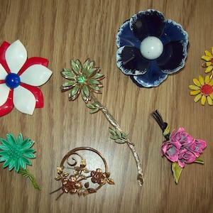 May include: A collection of vintage flower brooches in various colors, including red, white, blue, green, yellow, and pink. The brooches are made of metal and enamel, and feature different floral designs.
