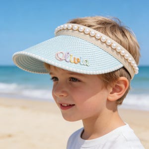 May include: A light blue visor with a woven texture, adorned with faux pearls along the brim and a beige band. The name "Olivia" is embroidered in colorful thread on the visor. The visor is designed for sun protection.