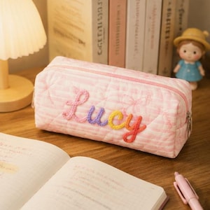 Op de afbeelding: Een rechthoekig gewatteerd etui met lichtroze en witte strepen en een subtiel strikjespatroon. De naam Lucy is op de voorzijde geborduurd in letters van roze, paars, geel en oranje garen, voorzien van een zilverkleurige metalen ritssluiting.