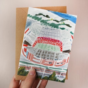 May include: Watercolor illustration of the Manchester United stadium on a greeting card. The stadium is depicted in red, white, and green, with the words "MANCHESTER UNITED" across the top. The card is held in front of a brown envelope.
