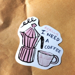 Puede incluir: Una taza de cerámica blanca con un asa negra y el texto "But First, Coffee" en negro, con una fuente de estilo manuscrito. La taza está sobre una superficie blanca.