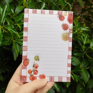 May include: A notepad with a red and white checkered border and lined pages. The notepad is decorated with watercolor illustrations of tomatoes in various stages of ripeness. The notepad is held in a hand.