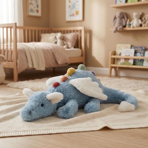 May include: A soft, blue dragon toy with white wings and colourful spine details. The stuffed animal is on a cream blanket in a child's bedroom. The dragon has a long tail and white accents on its ears. The room has a cot and bookshelves.