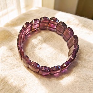 Puede incluir: Una pulsera elástica compuesta por numerosas cuentas de color púrpura translúcido, lisas y pulidas. Las cuentas son rectangulares con bordes redondeados y están dispuestas en una formación circular, reflejando la luz a través de sus superficies violetas.