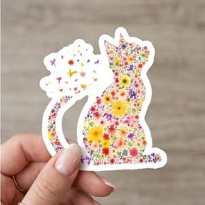 Peut inclure: Un autocollant blanc en forme de chat, orné de fleurs colorées. Le chat est assis, la queue enroulée. Les fleurs sont dans les tons de jaune, rouge, violet et blanc. L'autocollant est tenu par une main.