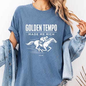 Peut inclure: T-shirt bleu avec l'inscription blanche "GOLDEN TEMPO MADE ME RICH". En dessous, un dessin blanc d'un cheval de course et de son jockey. Le jockey et le cheval portent le numéro "19". L'année "2026" est imprimée en dessous.