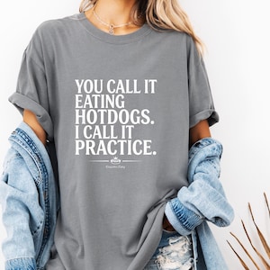 Puede incluir: Camiseta gris con el texto blanco: "YOU CALL IT EATING HOTDOGS. I CALL IT PRACTICE." Un pequeño diseño de corona y las palabras "Competitive Eating" están debajo. La camiseta es de estilo casual.