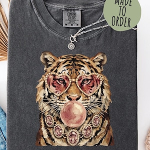 May include: A charcoal gray t-shirt featuring a graphic of a tiger wearing heart-shaped glasses and a jeweled necklace while blowing a pink bubblegum bubble. The shirt has a Comfort Colors label at the neck and a circular Made To Order graphic.