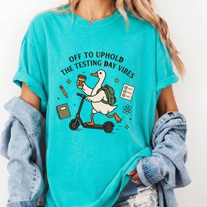 Peut inclure: T-shirt turquoise avec l'inscription « Off to Uphold the Testing Day Vibes ». Le motif représente un canard de dessin animé sur un scooter, tenant un café et portant un sac à dos. Autres graphiques : crayon, carnet et feuille de test.