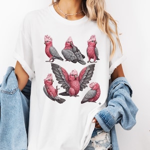 Puede incluir: Camiseta blanca con una ilustración en acuarela de cacatúas Galah rosas y grises. Las aves se muestran en diferentes poses, algunas con las alas extendidas. El diseño está centrado en la parte delantera de la camiseta.