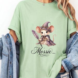 Peut inclure: T-shirt vert clair avec une illustration fantaisiste d'une souris portant un chapeau et une cape de sorcière, tenant une plume. Le nom "Mossie" est écrit en dessous en script.
