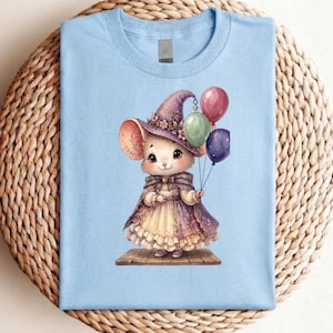 Peut inclure: T-shirt bleu clair avec une illustration fantaisiste d'une souris portant un chapeau et une cape de sorcière violets, tenant des ballons colorés. La souris est sur une plateforme en bois. Le design est détaillé et coloré.