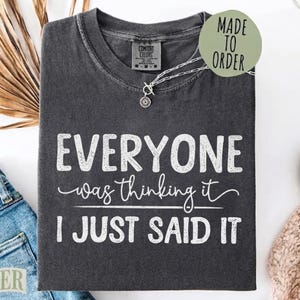 Peut inclure: Un t-shirt à col rond gris anthracite avec le texte blanc "EVERYONE was thinking it I JUST SAID IT" imprimé sur le devant. Le t-shirt présente un aspect délavé vintage et une étiquette de marque Comfort Colors à l'intérieur du col. Une chaîne argentée est posée sur le col.