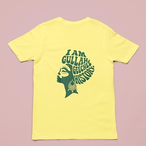 May include: A yellow t-shirt with a green graphic on the back. The graphic depicts a woman's profile and the text "I AM GULLAH GEECHEE HISTORY" in a stylized font. The woman is wearing earrings. The t-shirt is a short-sleeved style.