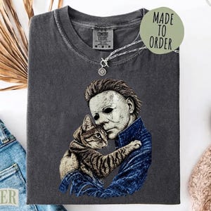 Peut inclure: Un t-shirt à col rond gris anthracite avec une impression graphique représentant un personnage au masque blanc portant une veste bleue et tenant un chat tigré brun. Le t-shirt porte une étiquette Comfort Colors et une étiquette Made To Order.