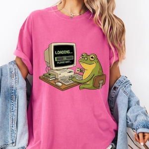 Peut inclure: T-shirt rose avec un dessin d'une grenouille devant un ordinateur. L'écran affiche "LOADING..." et "1% PLEASE WAIT...". La grenouille est verte et jaune, avec une tasse sur le bureau. Le t-shirt a des manches retroussées.