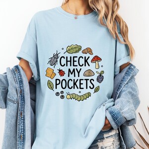 Peut inclure: T-shirt bleu clair avec l'inscription "CHECK MY POCKETS" entourée d'illustrations d'insectes, de feuilles et d'aliments. Le t-shirt est en tissu doux et à manches courtes.