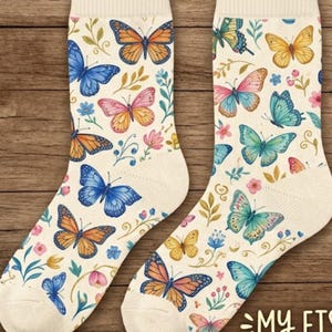 Op de afbeelding: Een paar crèmekleurige sokken met een kleurrijk vlinderpatroon. De sokken hebben verschillende vlinders in blauw, oranje, roze en geel, samen met bloemen- en bladaccenten. De sokken hebben een geribbelde boord.