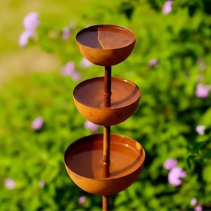 Può includere: Una vasca per uccelli in metallo color ruggine a tre livelli, dal design semplice ed elegante. Ogni ciotola è rotonda e sostenuta da un palo centrale. La vasca per uccelli è ambientata su uno sfondo sfocato di fogliame verde e fiori viola.