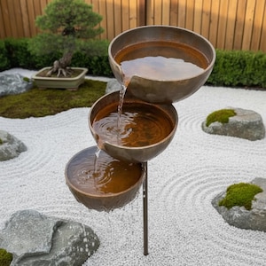 May include: A tiered water fountain featuring three stacked, circular bronze-toned bowls. Water flows from the top bowl into the middle bowl and then into the bottom bowl, creating a cascading effect. The fountain is supported by a thin metal pole.