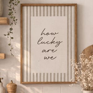 May include: A framed print with the text "how lucky are we" hangs above a wooden countertop. The kitchen scene includes a vase of dried flowers, a cutting board, and decorative bottles and jars. The color palette is neutral with natural wood tones.