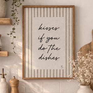 Puede incluir: Una impresión enmarcada con el texto "kisses if you do the dishes" en un entorno de cocina. La impresión tiene un fondo a rayas beige y blanco. Otros elementos de la cocina incluyen una tabla de cortar de madera, un jarrón con flores y una encimera de madera.