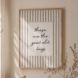 May include: A rectangular wall art print in a light wood frame featuring a vertical beige and white striped pattern with a central cream-coloured block containing the black cursive text that reads, these are the good old days.