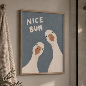 Può includere: Una stampa artistica da parete rettangolare in una cornice di legno chiaro che raffigura due oche bianche con becchi arancioni su uno sfondo blu tenue. Il testo NICE BUM è stampato in bianco, con un carattere scritto a mano nell'angolo in alto.