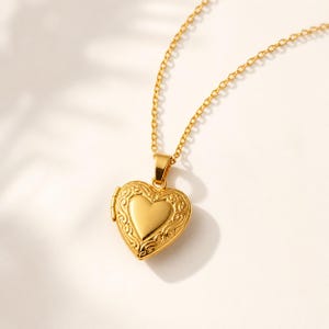 Könnte beinhalten: Goldfarbene Halskette mit herzförmigem Medaillon. Das Medaillon hat ein verziertes Design an den Rändern und ein glattes Herz in der Mitte. Die Halskette hat eine feine, goldfarbene Kette.