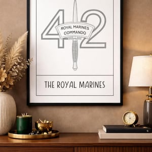 May include: A framed print featuring the text "42 ROYAL MARINES COMMANDO" in gray, with a dagger illustration. The words "THE ROYAL MARINES" are at the bottom. The print is in a black frame, displayed on a wooden surface.