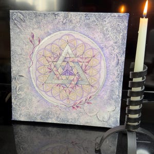 Op de afbeelding: Vierkante canvas kunst met een getextureerde, lichtpaarse achtergrond. Het kunstwerk toont een geometrisch ontwerp met een zilveren driehoek en bloemmotieven in paarse en gele tinten. Een brandende kaars in een zwarte metalen houder staat rechts.