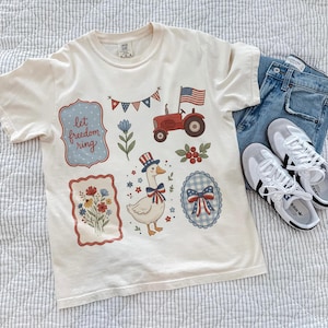 Può includere: T-shirt color crema con grafica patriottica, tra cui un trattore rosso, una bandiera americana e un'oca con un cappello. La maglietta è abbinata a jeans blu e scarpe da ginnastica bianche.