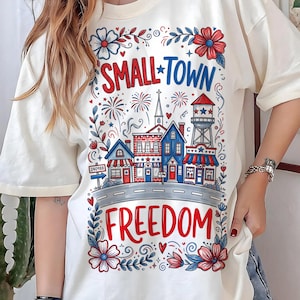 Könnte beinhalten: Cremefarbenes T-Shirt mit patriotischem Design. Die Grafik zeigt die Worte "SMALL TOWN FREEDOM" und eine Stadtansicht mit Gebäuden, Feuerwerk und floralen Akzenten in Rot, Weiß und Blau. Ein Straßenschild zeigt "240988."