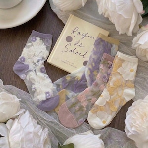 May include: Sheer socks with floral embroidery in shades of white, purple, and yellow are displayed with a book titled "Rayon de Soleil." The socks have different colored toes and cuffs. The background includes white flowers and a white sheer fabric.