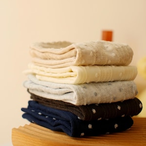 May include: A stack of five pairs of folded socks in various colors including beige, cream, light gray, dark brown, and navy blue. Each pair features a subtle textured dot pattern and vertical ribbed detailing along the fabric surface.