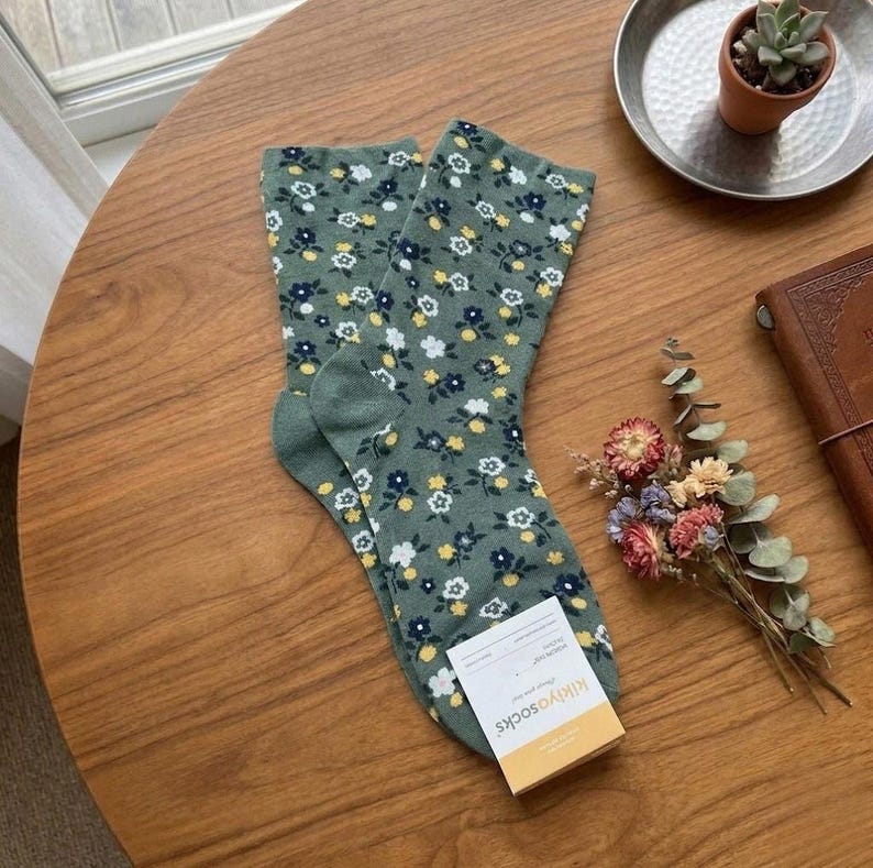 Pode incluir: Um par de meias verdes com um padr&atilde;o floral com flores brancas, amarelas e azul marinho. As meias est&atilde;o numa superf&iacute;cie de madeira, com uma etiqueta "kikiyosocks".