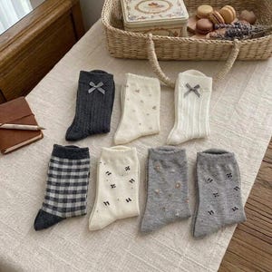 Può includere: Sette paia di calzini di media lunghezza in vari colori e fantasie, tra cui grigio antracite tinta unita con fiocco, crema con stampe floreali, bianco con trama a trecce e fiocco, quadretti vichy bianchi e neri, crema con piccoli fiocchi.