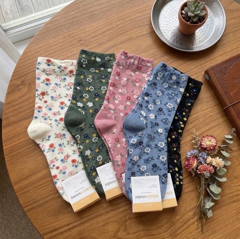 Pode incluir: Uma cole&ccedil;&atilde;o de cinco pares de meias com padr&otilde;es florais em v&aacute;rias cores, incluindo branco, verde, rosa, azul e preto. Cada meia apresenta um design floral diferente. As meias s&atilde;o exibidas numa superf&iacute;cie de madeira.