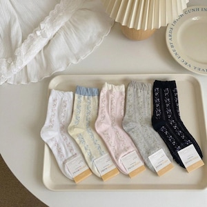 May include: A collection of six pairs of patterned socks in various colors, including white, pale yellow, pink, gray, and navy. Each sock features a floral design and is displayed on a white tray. The socks have tags attached.