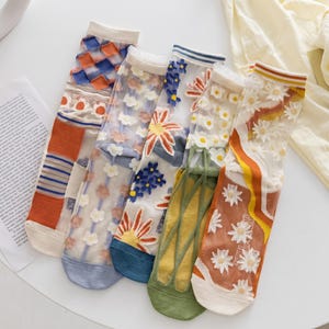 May include: Five pairs of sheer crew socks featuring varied designs. Patterns include blue and orange checkered blocks, white floral embroidery, yellow and orange sun motifs, green leaf-like structures, and white daisy appliques on orange.