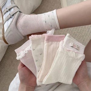 May include: A collection of four pairs of ankle socks in shades of cream, pale pink, and white. The socks feature varied textures including ribbed fabric, delicate lace trim, small floral patterns, and a small rectangular tag with floral print.