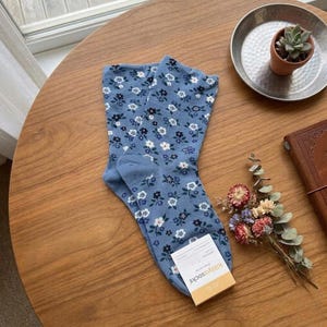 Pode incluir: Um par de meias azul claro com um padr&atilde;o floral de flores brancas e azul escuro. As meias est&atilde;o numa mesa de madeira. Uma pequena etiqueta diz "killyasocks".
