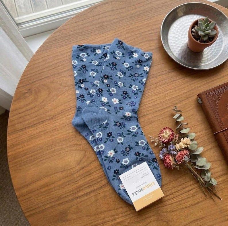 Pode incluir: Um par de meias azul claro com um padr&atilde;o floral de flores brancas e azul escuro. As meias est&atilde;o numa mesa de madeira. Uma pequena etiqueta diz "killyasocks".