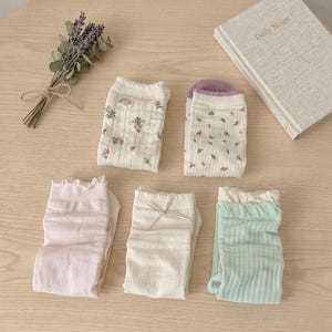 May include: An assortment of five pairs of ankle socks in various pastel colors, including pink, white, and mint green. Some socks feature floral patterns and delicate lace details. A small bundle of dried lavender and a notebook with the text "Daily Notes" are also present.
