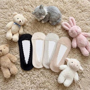 May include: A collection of no-show socks in black, beige, and light pink, with a lace trim. The socks are surrounded by plush toys, including teddy bears, a kitten, a bunny, and a lamb, all on a textured, light brown surface.