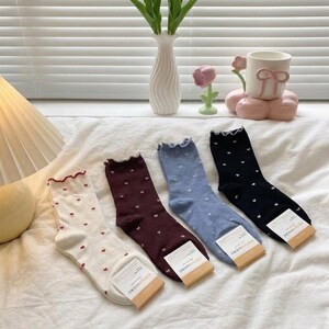 May include: Four pairs of socks in cream, burgundy, light blue, and black, each adorned with tiny white hearts. The socks have a ruffled trim at the top and small tags at the bottom. The socks are displayed on a white surface.