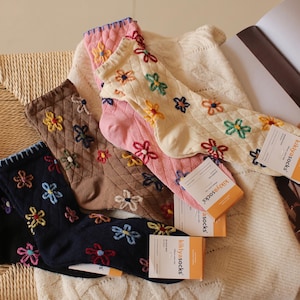 Peut inclure: Une collection de quatre paires de chaussettes montantes en noir, marron, rose et crème, chacune présentant une texture en losanges et des motifs de fleurs brodées multicolores. Chaque paire inclut une étiquette blanche et orange kikiyasocks.