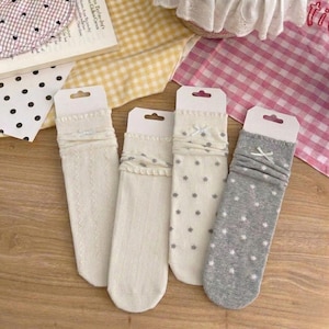 May include: Four pairs of knee-high socks in various designs. Two pairs are cream-colored, one with a textured pattern and the other with a ruffled top. The other two pairs are white with gray star patterns and gray with white star patterns.